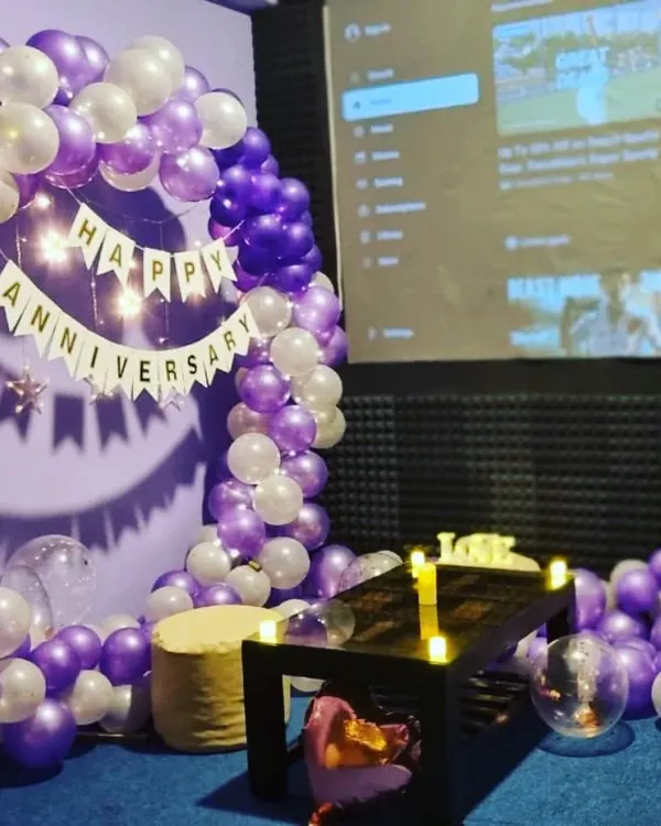 A beautiful anniversary setup with a purple and white balloon arch and a 'Happy Anniversary' banner. The soft lighting from the fairy lights adds a romantic touch to the room.