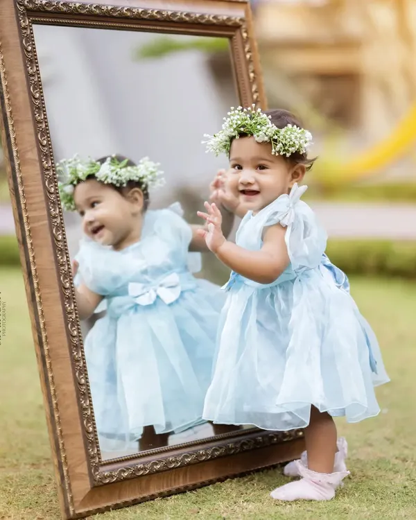 An adorable baby girl in a light blue dress and a floral crown looks at her reflection in a large, ornate mirror during an outdoor photoshoot. This creative setup makes for a magical portrait.