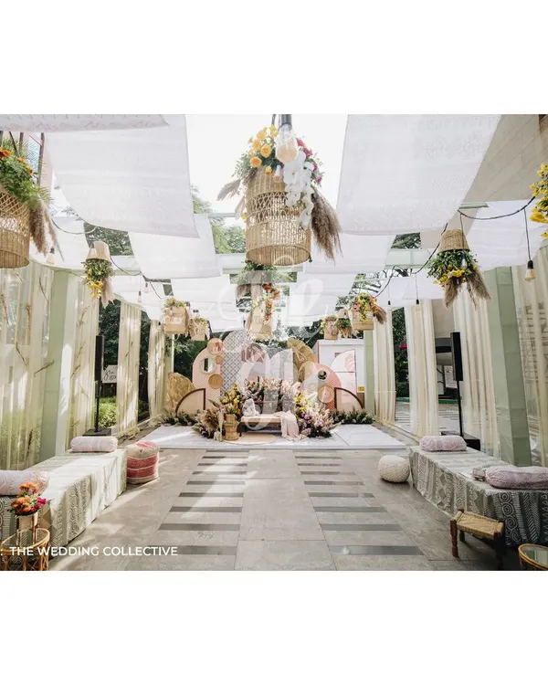 A wide view of our boho-pastel Mehendi setup, designed for a relaxed sundowner celebration. The space features low lounge seating, printed drapes, and hanging baskets filled with flowers, creating a comfortable and chic atmosphere.