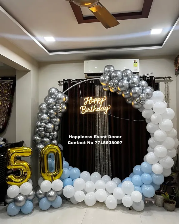 A stylish 50th birthday home decoration. This setup features a balloon half-arch in silver, white, and blue, a 'Happy Birthday' neon sign, and large foil balloons for the number '50'.