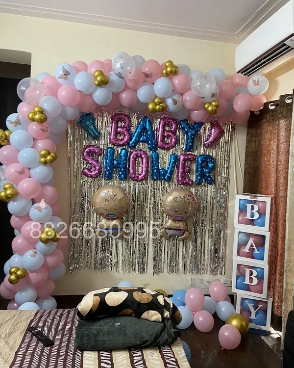 A complete baby shower decoration for a home celebration. The setup includes a balloon arch in pink, light blue, and gold, a silver fringe backdrop, and 'BABY' blocks on the side.