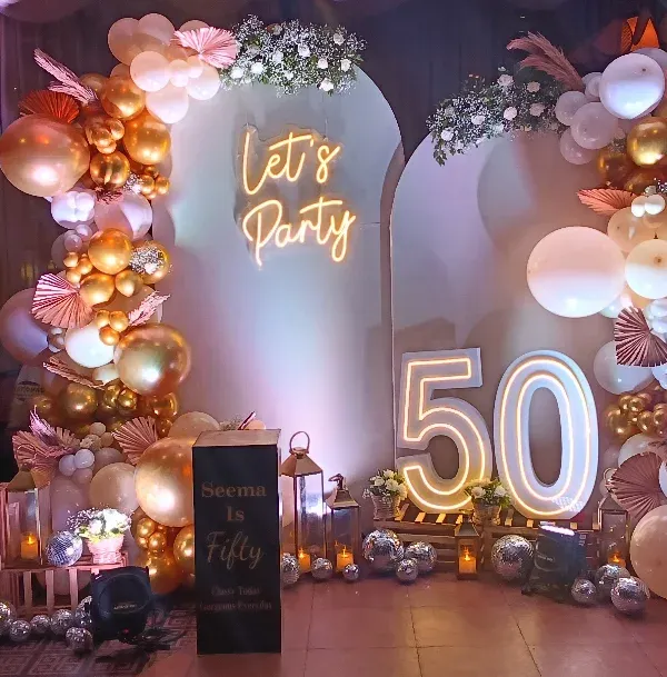 A fabulous 50th birthday celebration! This setup features a "Let's Party" neon sign, large marquee numbers, and a stunning balloon arrangement in rose gold, white, and gold.
