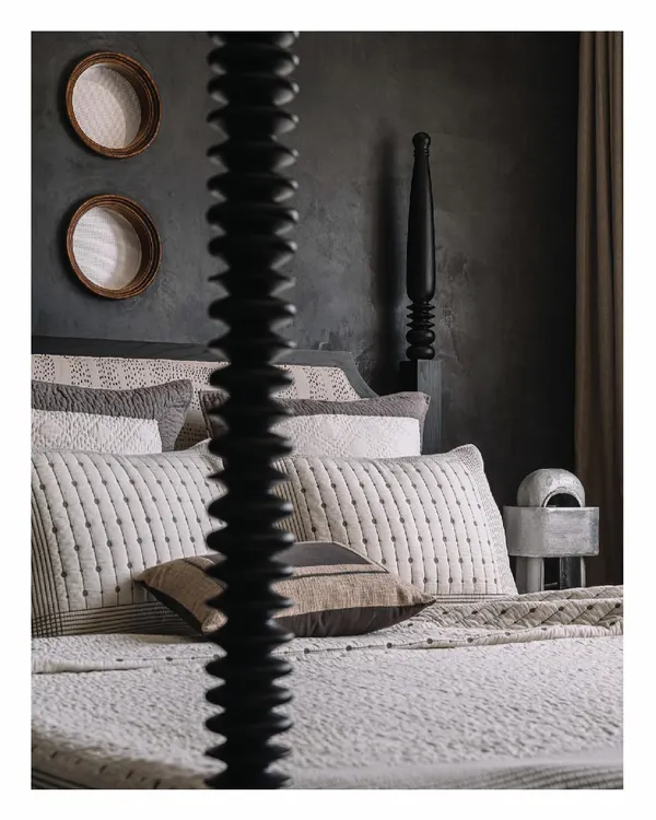 The master bedroom in our 'Aria' project uses a stark, masculine palette of black and white with bursts of mud red. A custom black burnt-wood four-poster bed creates a dramatic centerpiece, viewed here through its intricately turned post, adding a layer of sculptural detail.