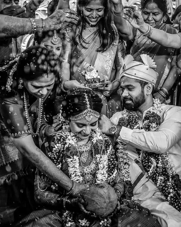 This black and white image captures the intensity and joy of a wedding ritual. The high contrast and surrounding family members create a powerful, timeless scene focused on the couple.