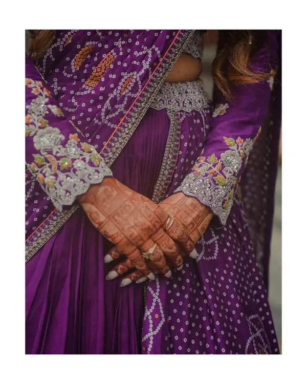 A close-up on the exquisite details of the bride's purple bandhani-style lehenga. The intricate embroidery and delicate henna on her hands tell a story of tradition and craftsmanship.
