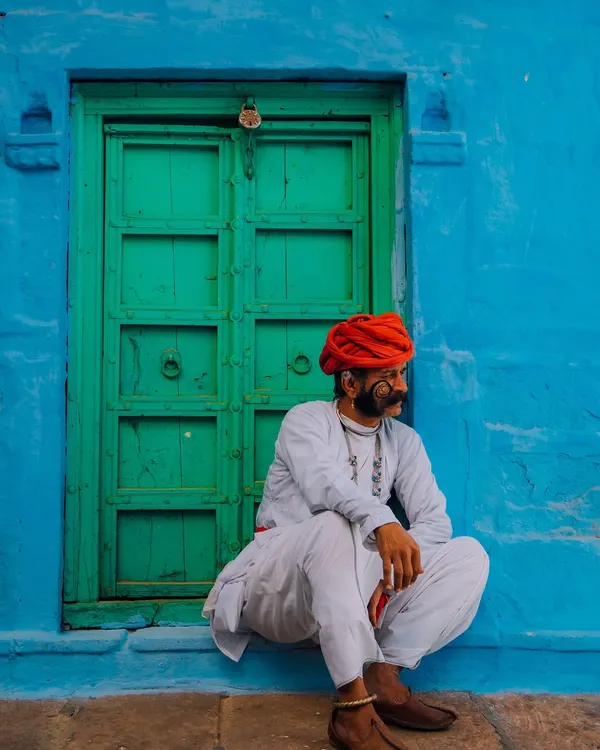 A man with a magnificent mustache and a red turban sits against a vibrant blue wall with a green door in Jodhpur. This is the quintessential color palette of Rajasthan, bold and full of character.