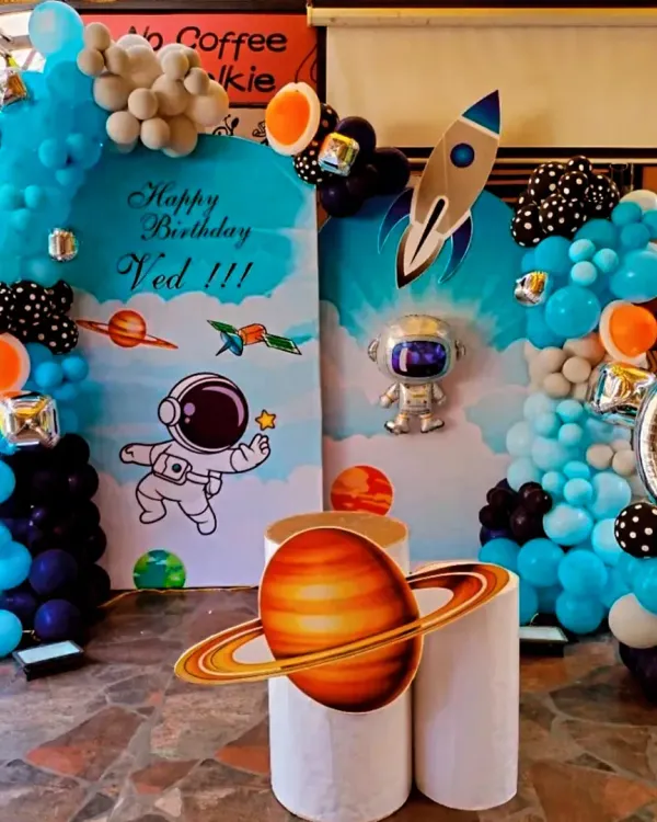 An out-of-this-world space-themed birthday party for Ved. This setup includes a large balloon arch in shades of blue, astronaut and rocket cutouts, planet props, and a personalized backdrop to create an exciting adventure for the birthday boy.