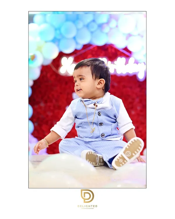 An adorable portrait of a one-year-old boy in a cute blue vest, sitting happily amidst balloons at his first birthday party.