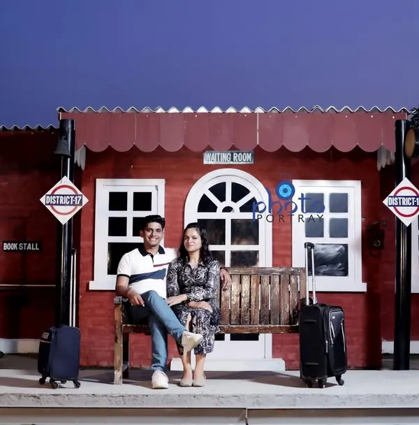 A travel-themed pre-wedding shoot using the vintage railway station set. The couple is dressed in casual attire, sitting on a bench with luggage props to complete the scene.
