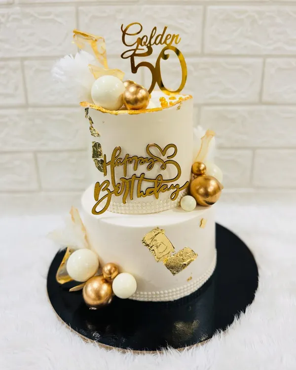 Celebrating a golden 50th birthday with this stunning two-tiered cake. It's decorated with white and gold spheres, gold leaf, and elegant toppers for a truly luxurious feel.