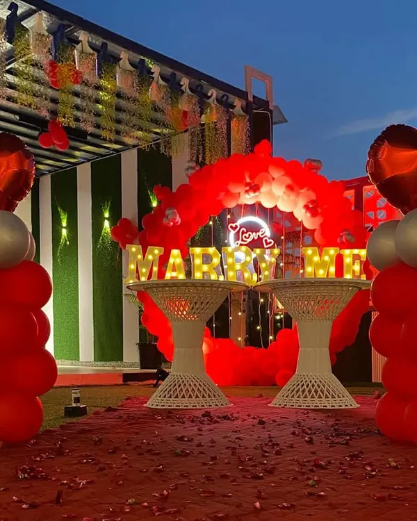 A beautiful daytime view of a rooftop proposal decoration. The setup includes bold red balloon pillars with heart toppers, a red carpet aisle, and large illuminated "MARRY ME" letters, all ready for that magical moment.
