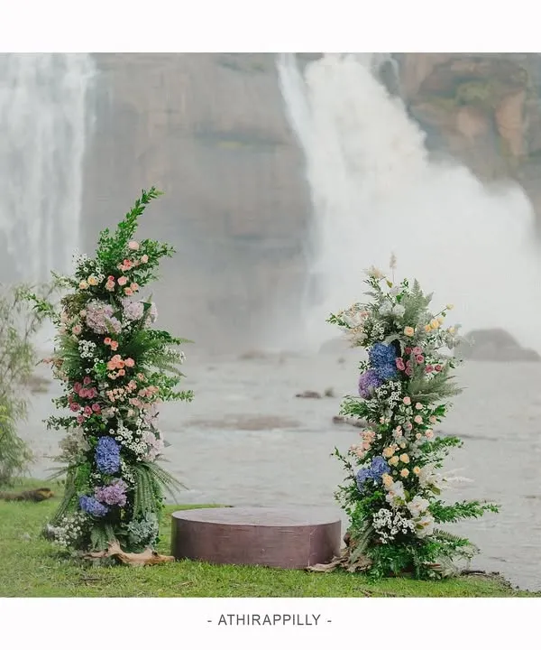 The stage is set for a vow renewal ceremony at the majestic Athirapilly waterfalls. The simple, elegant decor with floral pillars stands in beautiful contrast to the raw power of nature, creating a truly unique wedding venue.