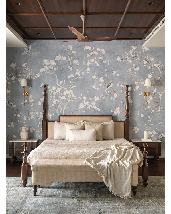 The master bedroom at Tinted Tapestries is a dreamy, tranquil sanctuary. The focal point is a custom four-poster bed in dark wood, set against an exquisite wallpaper adorned with delicate floral and avian motifs.