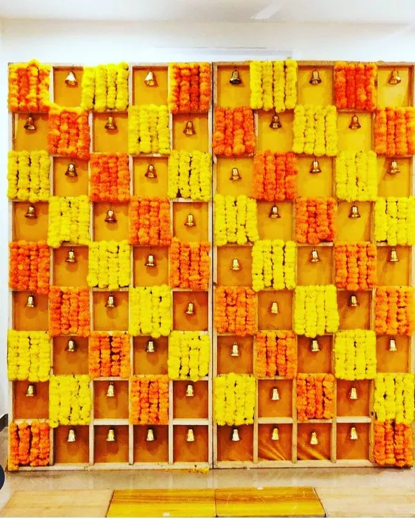 This unique backdrop is constructed from a wooden grid, with each section holding marigold flowers and a small brass bell. It's a visually stunning design that adds a beautiful sound element to the celebration.