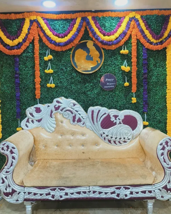 A classic stage setup for a traditional baby shower, featuring an ornate sofa against a green grass wall backdrop with marigold flower garlands.