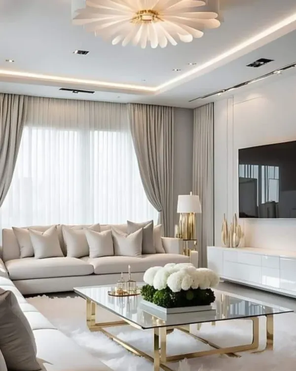 This living room design emphasizes serene elegance with a neutral color palette of cream and beige. A large L-shaped sofa offers plush comfort, while the gold-accented glass coffee table and a unique floral-inspired ceiling light add a touch of glamour.