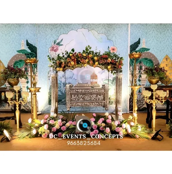 A grand and traditional cradling ceremony setup with a peacock-themed backdrop. The decor includes an ornate cradle, traditional brass lamps (Samai), and beautiful floral arrangements.