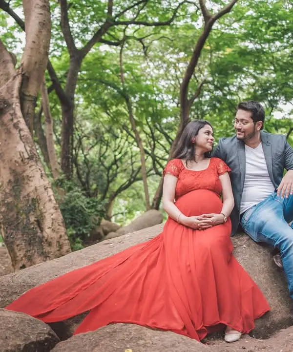 A serene moment between an expecting couple, sitting on a large rock formation in a park, surrounded by greenery. The flowing red gown adds a beautiful pop of color.