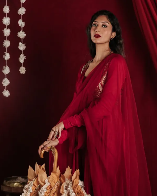 This studio shot focuses on the interaction between the model and the designer umbrella. The pose is directed to highlight the product's unique shape and handcrafted details, set against a rich, deep red that complements the model's attire.