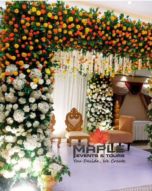 A beautiful mandap setup for a wedding ceremony, featuring pillars wrapped in white and marigold flowers. The traditional seating for the bride and groom is set for the sacred rituals to begin.