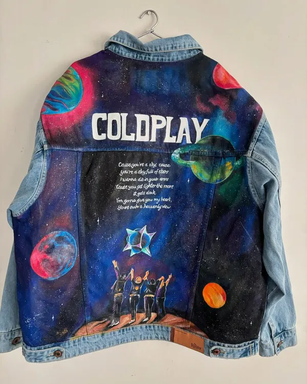This custom-painted denim jacket was created for a client attending a Coldplay concert. The entire back panel is a canvas, featuring a vibrant galaxy scene, the band's silhouette, and iconic lyrics from "A Sky Full of Stars," making it the ultimate piece of fan merchandise.