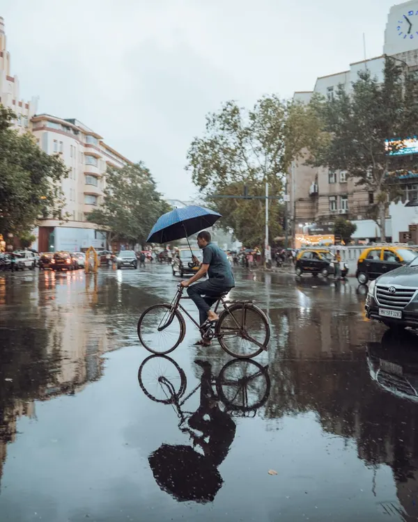 A man cycles through a waterlogged Mumbai street during the monsoon, holding an umbrella to balance against the rain. This shot perfectly encapsulates the 'Mumbai grit' and the resilience of its people.