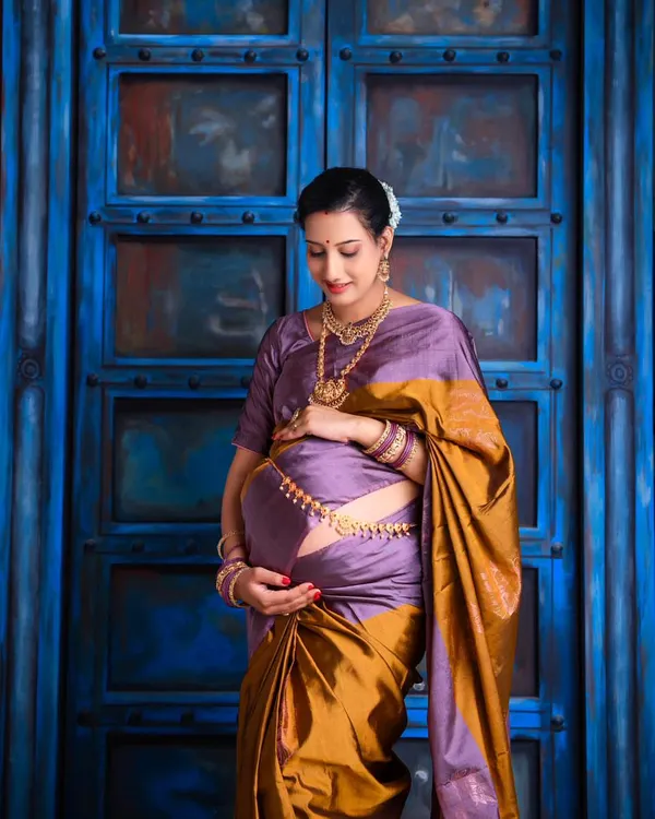 A beautiful solo portrait of the mom-to-be in a traditional purple and gold silk saree. She cradles her bump, radiating grace and serenity against a rustic blue door.