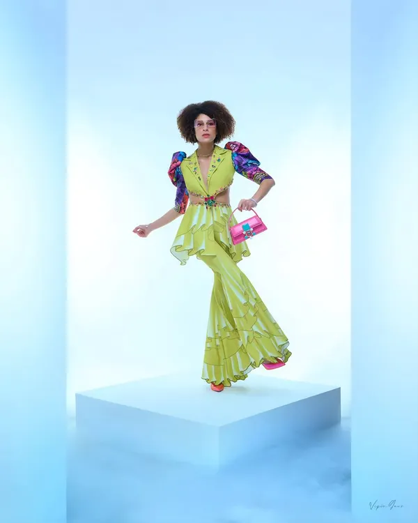 A designer fashion shoot showcasing a vibrant lime green co-ord set. The model's pose is dynamic, highlighting the ruffled details and unique silhouette of the outfit against a clean, ethereal blue-and-white backdrop.