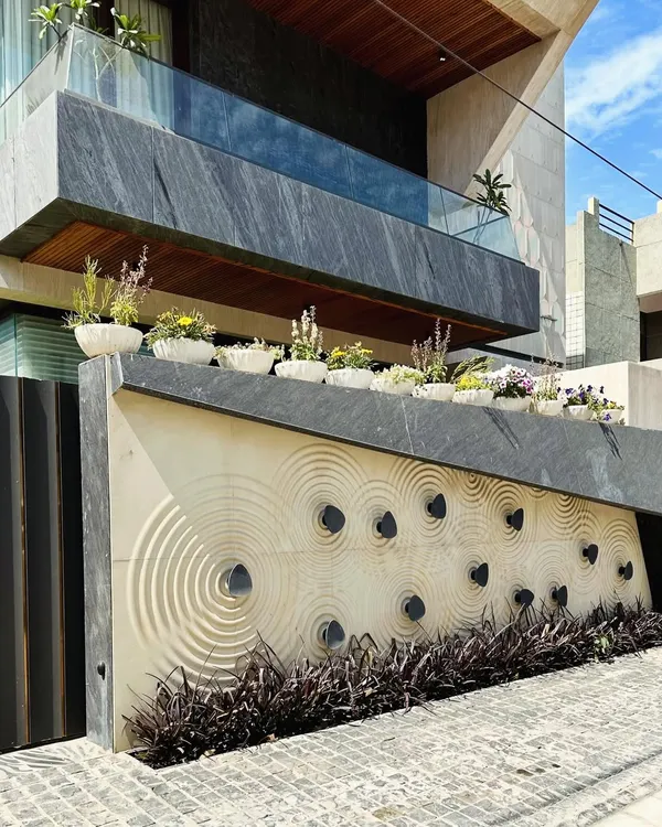 A stunning architectural detail. This boundary wall in Faridabad features a custom CNC-carved stone panel with a concentric circle pattern, topped with a row of flowering plants in matching pots.