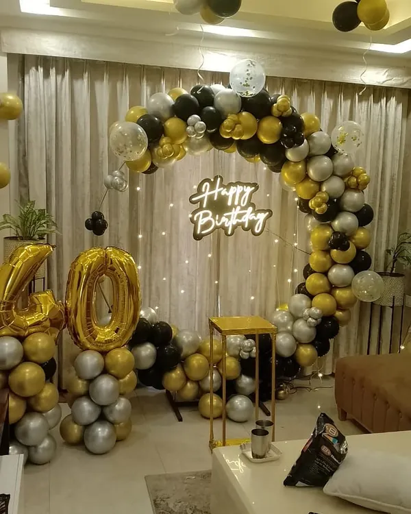 A stylish 40th birthday celebration at home. This setup features a circular balloon arch in black, gold, and silver, a 'Happy Birthday' neon sign, and large '40' foil balloons.