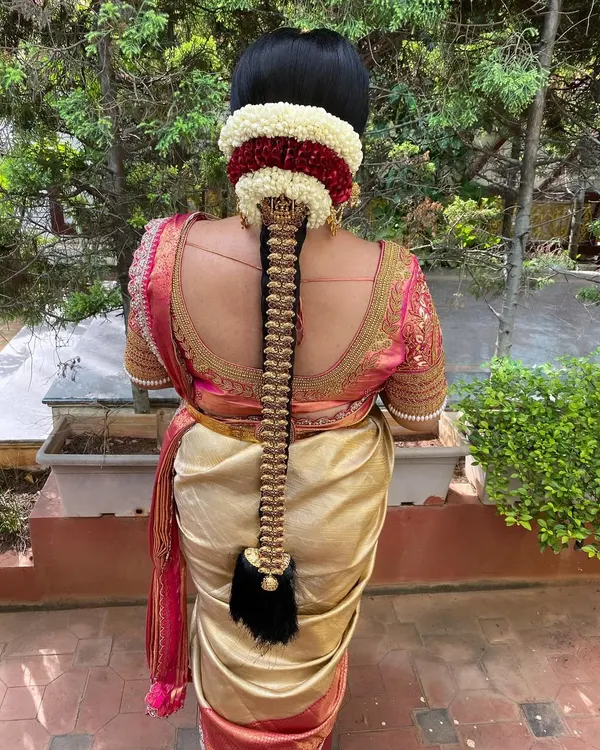 A classic Muhurtham look featuring a long, neat braid decorated with a full set of traditional gold temple jewellery. The base is adorned with a layer of jasmine and red flowers for a vibrant, customary finish.
