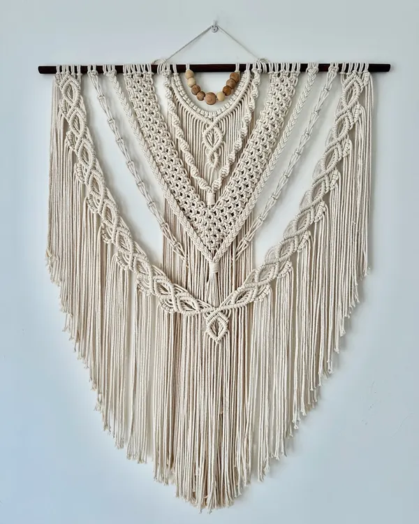 This large, V-shaped macrame wall hanging is made entirely with natural off-white cord and features wooden beads for an earthy touch. The design includes multiple layers of intricate knots, creating amazing texture and a classic bohemian look for any large wall.
