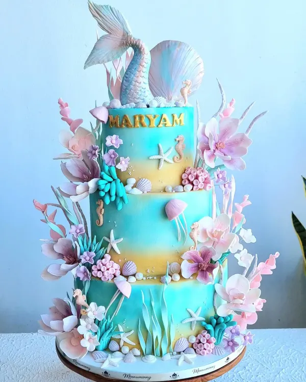 This three-tier mermaid theme cake is a perfect example of a magical underwater world brought to life. It features a buttercream base with a soft color gradient, detailed with handcrafted fondant elements like a mermaid tail, seashells, corals, and seahorses for a truly enchanting birthday centerpiece.