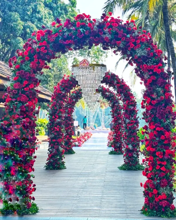 A grand entrance for a destination wedding, featuring a series of arches covered in lush red and pink roses, with crystal chandeliers hanging within each arch to create a luxurious walkway.