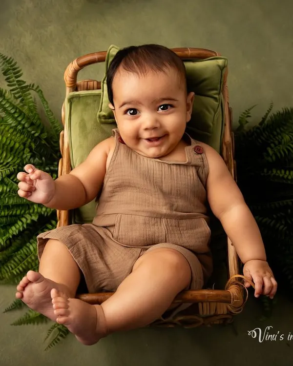 A cheerful smile from a little one. This infant portrait, taken between 2 to 5 months, captures a happy baby sitting in a wicker chair surrounded by green ferns.