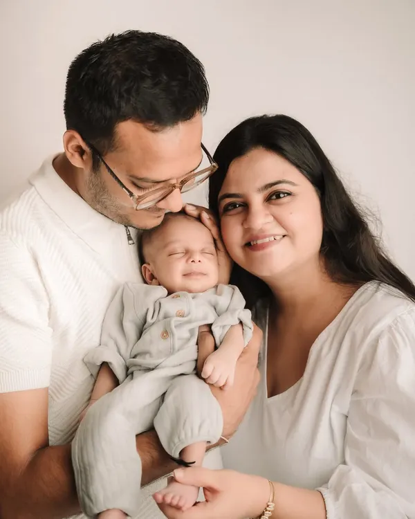 A perfect family portrait with a smiling newborn held lovingly by his parents. The simple white background keeps the focus on them.