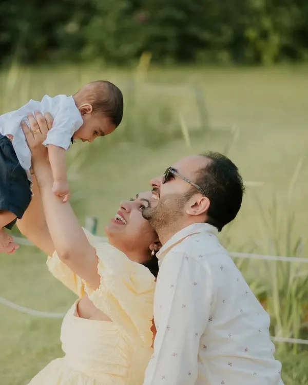 A joyful moment of a mother lifting her baby into the air during an outdoor family photoshoot. The natural light and green surroundings create a fresh and happy scene.