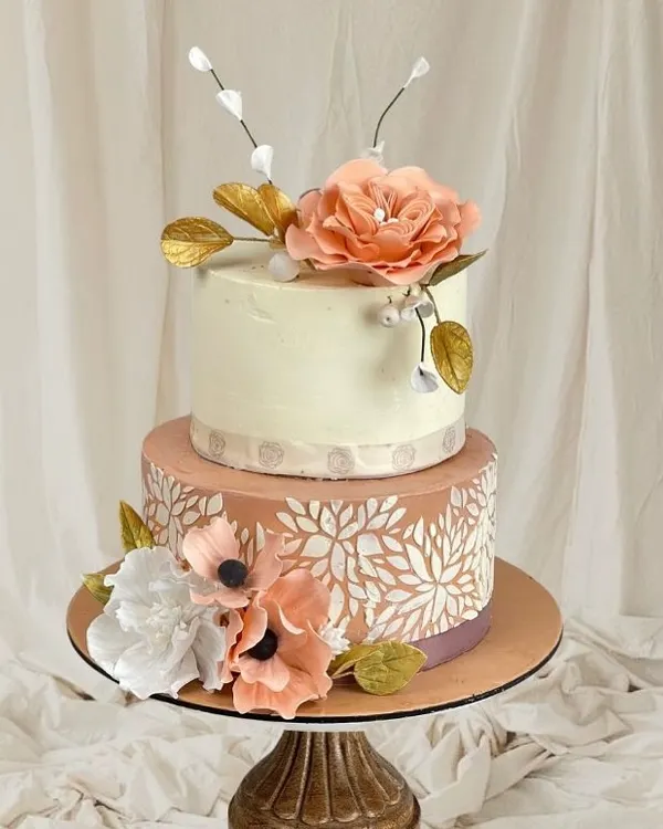 This two-tiered cake is a study in elegance, combining delicate stencil work on the bottom tier with beautiful, handcrafted sugar flowers on top.