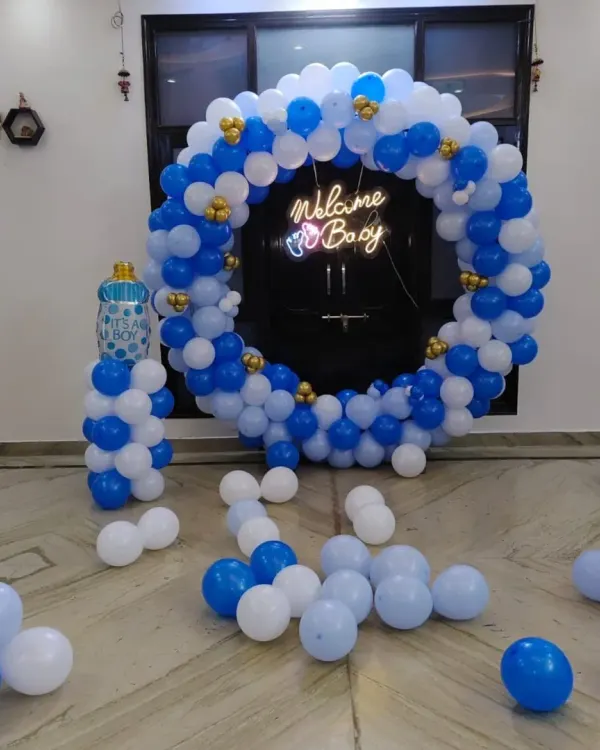 A complete "Welcome Boy" decoration in a living room. This setup includes a circular balloon arch with a neon sign, loose balloons on the floor, and a balloon pillar with a baby bottle foil balloon.