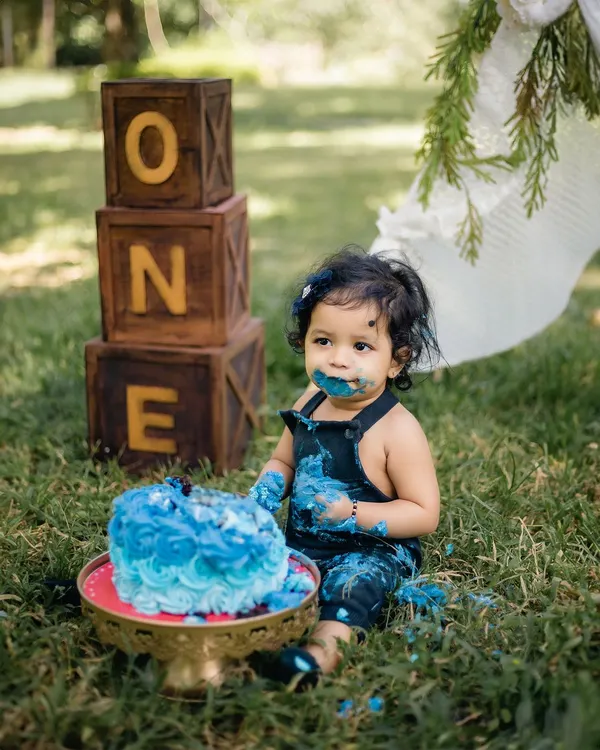 A one-year-old gets delightfully messy with a blue birthday cake during an outdoor cake smash session. The rustic "ONE" blocks and teepee in the background create a perfect boho-chic setup.