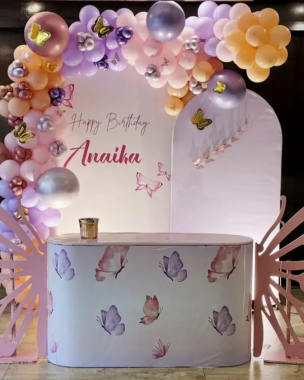 A magical butterfly theme for Anaika's birthday. The setup includes a custom-printed cake table, large pink butterfly wing cutouts, and a pastel balloon arch with delicate butterfly accents.