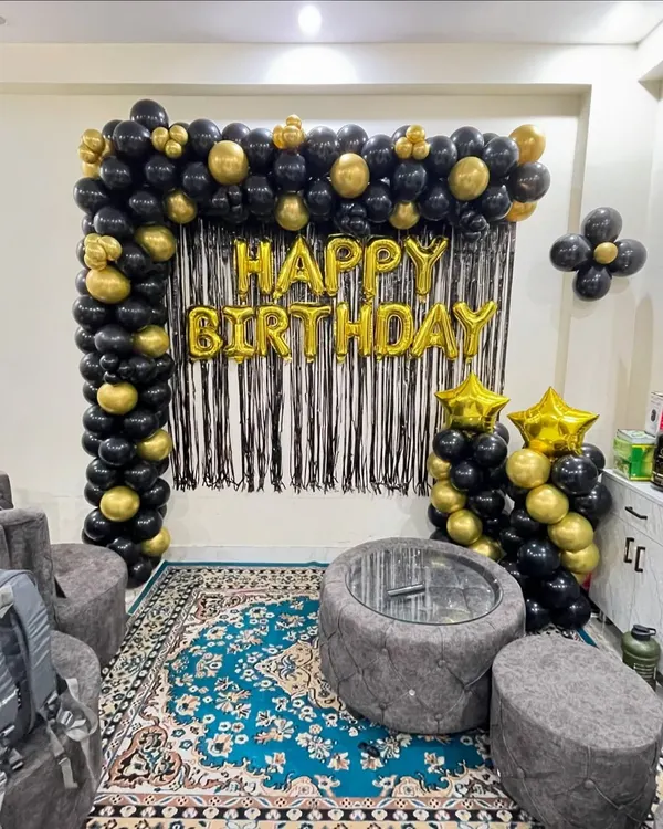 A classic and elegant birthday decoration for a living room. This setup features a black and gold balloon arch over a black fringe backdrop, with "Happy Birthday" foil letters.