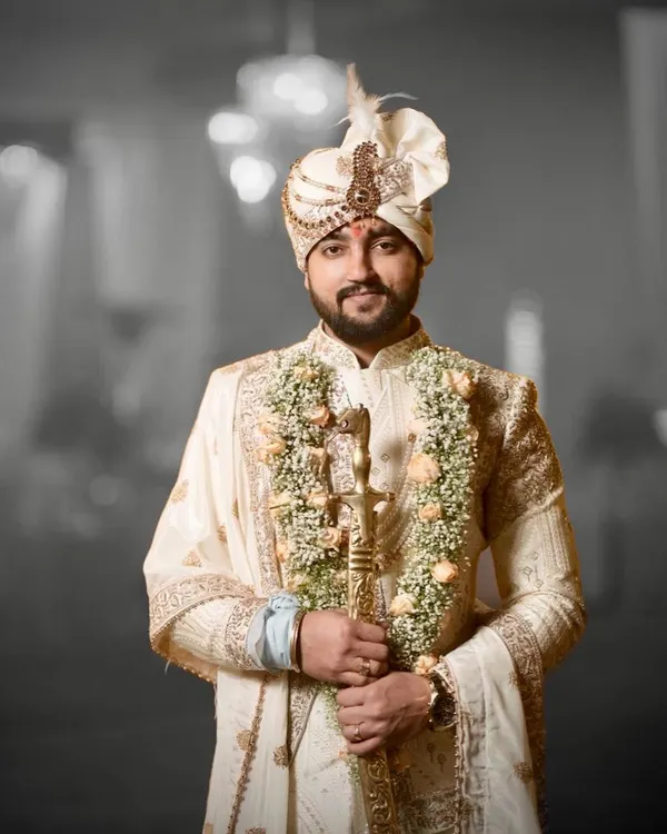 A groom ready for his ceremony, wearing a classic ivory sherwani. The outfit is detailed with fine embroidery and paired with a traditional safa and a ceremonial sword, completing the royal look.