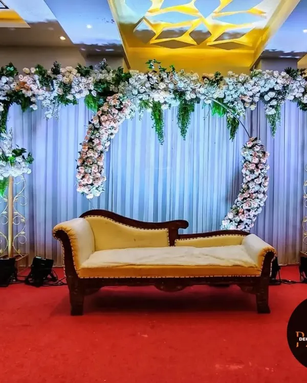 This elegant stage decoration for a ring ceremony features a beautiful arch of white and blush pink flowers. A classic chaise lounge provides sophisticated seating for the couple against a simple white curtain backdrop.