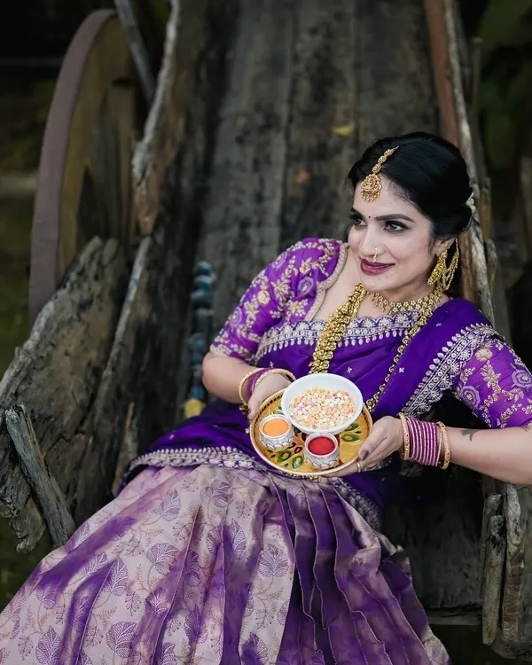 A beautiful portrait for Sankranti, with the model in a traditional purple half saree, holding a plate of offerings. The rustic bullock cart adds a classic touch.