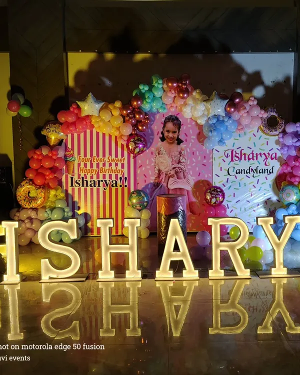 This 'Candyland' themed birthday party features large, light-up marquee letters spelling 'AISHARYA'. The backdrop includes a colorful organic balloon garland with donut and candy accents to bring the sweet theme to life.