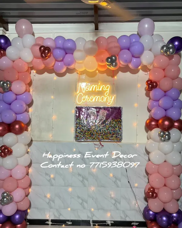 This is a popular decoration for a naming ceremony at home. It features a full balloon arch in multiple colors with a 'Naming Ceremony' neon sign and a name reveal board against a simple tiled wall.