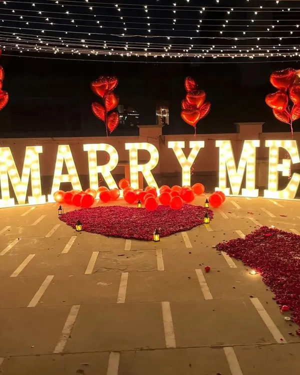 A grand rooftop proposal setup in Bengaluru featuring large, illuminated 'MARRY ME' marquee letters. We created a romantic pathway and heart shape using fresh red rose petals, complemented by heart-shaped foil balloons and soft candlelight for an unforgettable moment.