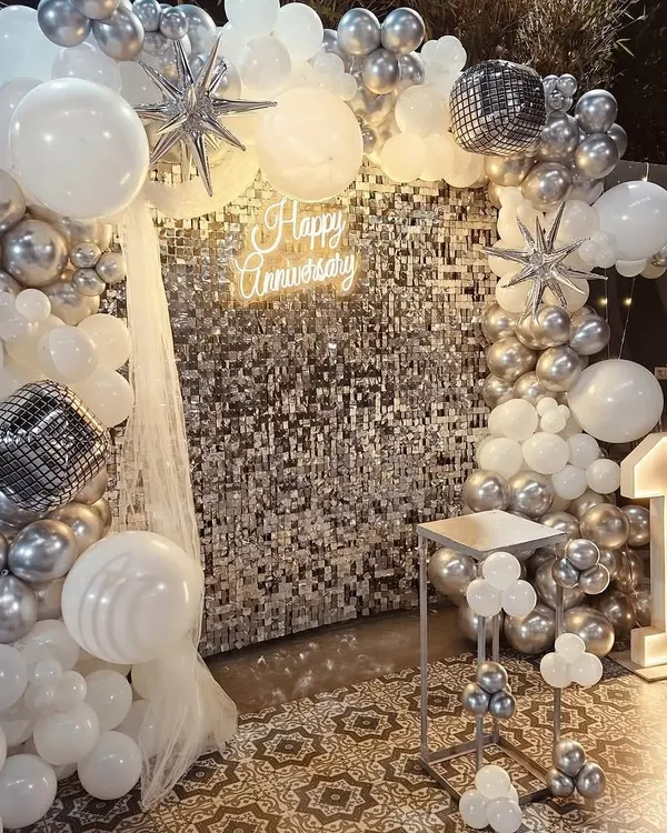 A glamorous first-anniversary party with a silver shimmer wall, a "Happy Anniversary" neon sign, and a silver and white balloon arch. The light-up number '1' marks the special milestone.