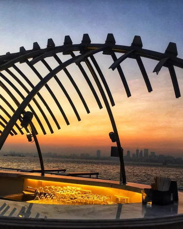 The open-air bar of the Queensline Sea Yah floating restaurant, silhouetted against the Mumbai skyline at sunset. This sculptural element, inspired by a whale skeleton, serves as the centerpiece of the upper deck, offering panoramic sea views.